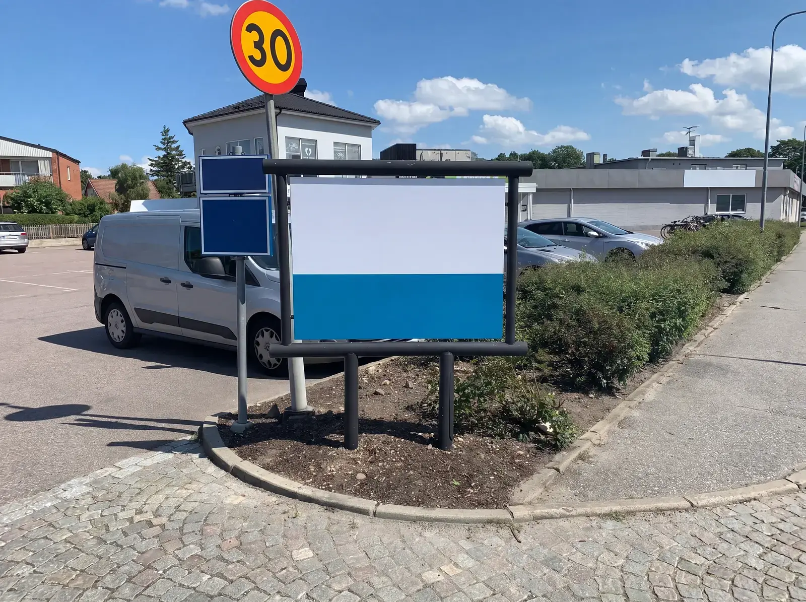 Informationstavla och hastighetsskylt vid parkeringsplats med grönska och byggnader i bakgrunden.